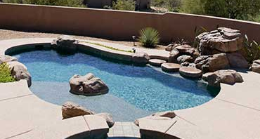 A freeform swimming pool on a concrete patio, with a large boulder in the water and rock formations along the edge.