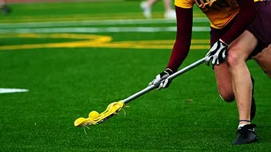 Gloved lacrosse player scoops a yellow ball from green turf.