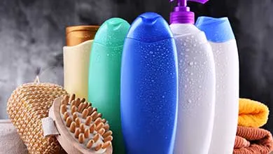 Assorted bath products: colorful shampoo and soap bottles with water droplets, a wooden body brush, loofah, and folded towels.