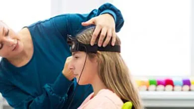 A technician in a blue shirt fits a head-mounted sensor onto a seated woman with blonde hair.