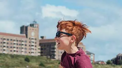Profile of a smiling boy with windswept red hair and sunglasses; a large building and partly cloudy sky in the background.