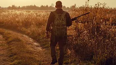 Man with backpack and rifle walks dirt path through tall grass at sunrise.