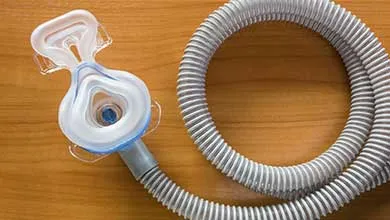A CPAP mask with a clear cushion is connected to a gray, corrugated hose, both lying on a wooden surface.