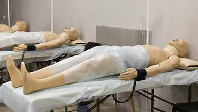 Two medical training mannequins lie on separate cots, partially covered with white sheets, with black straps attached to their wrists.