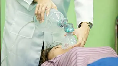 A healthcare worker in a white coat gives oxygen to a patient lying down using a bag valve mask.