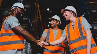 Workers wearing safety vests and hard hats shaking hands inside a warehouse environment.