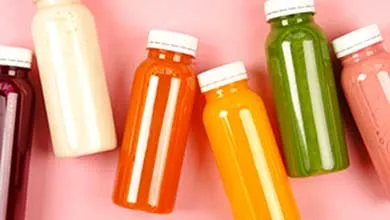 Overhead view of clear plastic bottles filled with orange, green, pink, and white juices, arranged on a pink background.