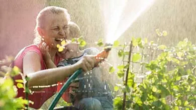 A woman and a child laugh while playing with a water hose in a sunny garden.