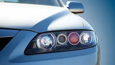 Close-up of a silver car’s front headlight assembly with multiple circular lights, including the main headlight and an amber turn signal, against a clear blue sky.