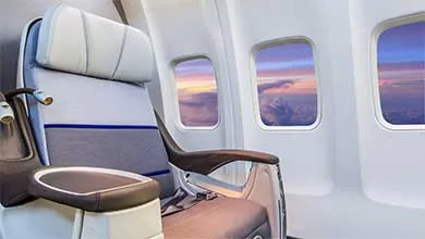 A light blue and gray airplane seat with a dark armrest sits beside three windows showing a vibrant sunset sky in pink, purple, and orange above scattered clouds.