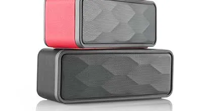 Two rectangular portable Bluetooth speakers stacked vertically on a white background. The top speaker has bright pink side panels and a dark gray front grille with a diamond-shaped geometric pattern. The bottom speaker is entirely dark gray or black with a matching textured diamond-patterned grille.